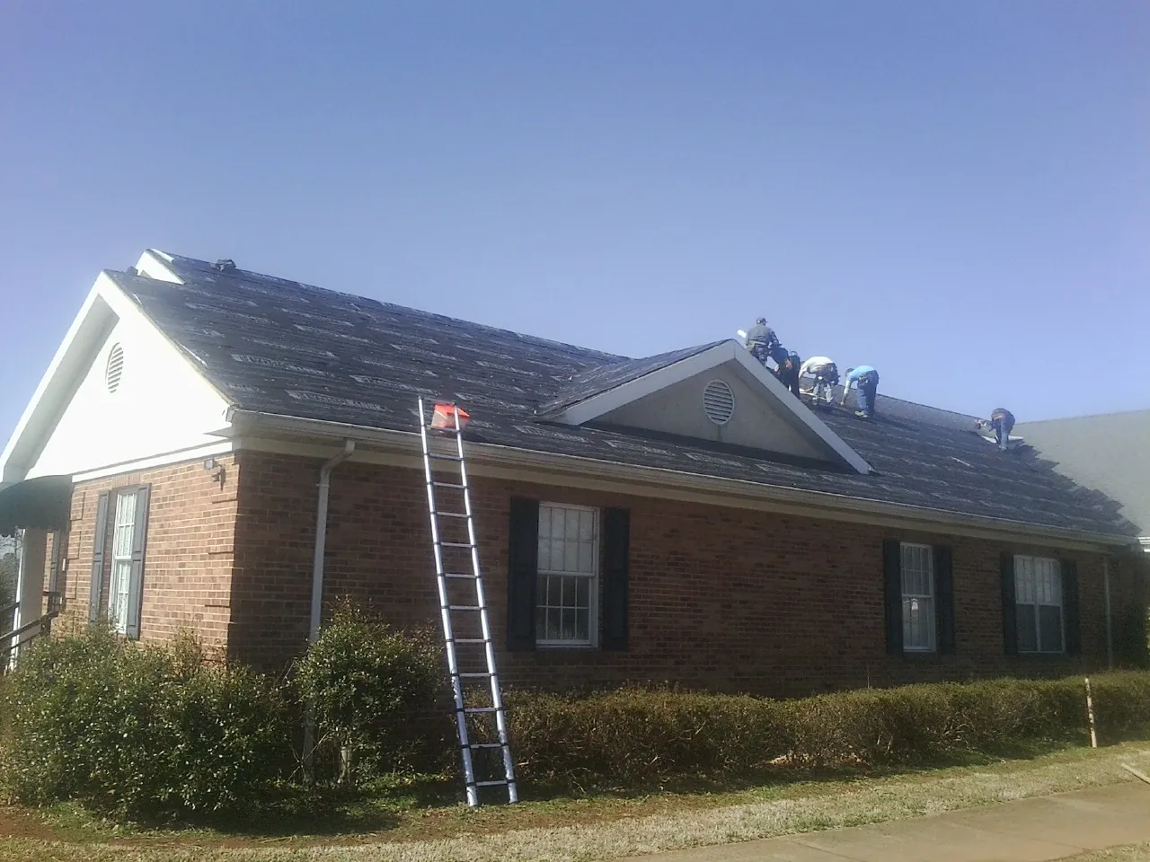 Crew tearing shingles off a brick ranch roof with a ladder set — Metal Roof Repair work by our crew in Clyo