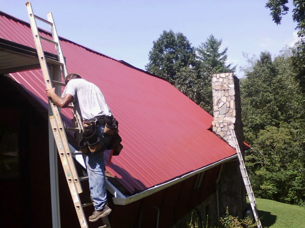 Roofer on a ladder beside a red metal roof and stone chimney — Soffit & Fascia Repair by Roofing Brothers in Clyo